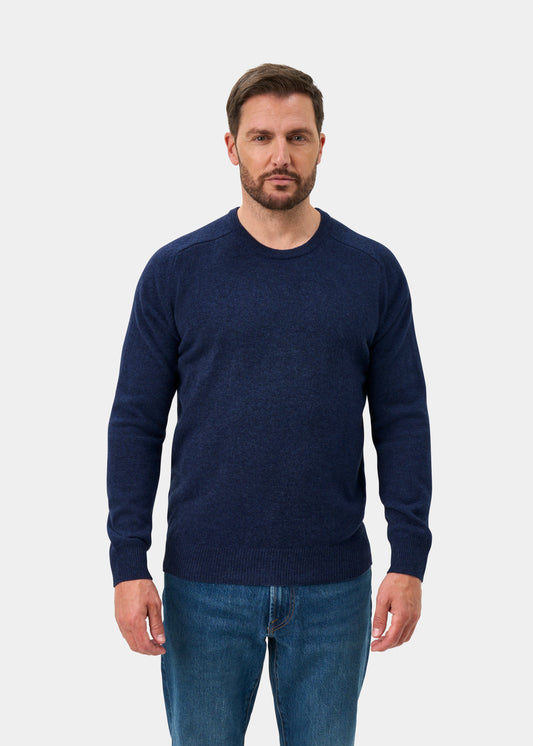 A man wears the Dorset Lambswool Crew Neck Jumper in Rhapsody, classic fit, with blue jeans against a light gray backdrop.