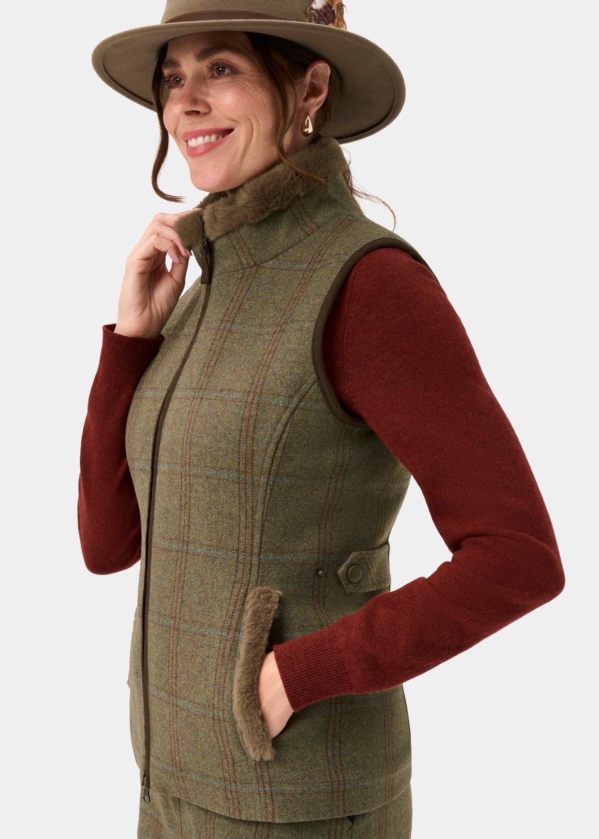 Smiling woman in Combrook Ladies Tweed Gilet in Laurel, red sweater, tan hat, looking to the side on white background.