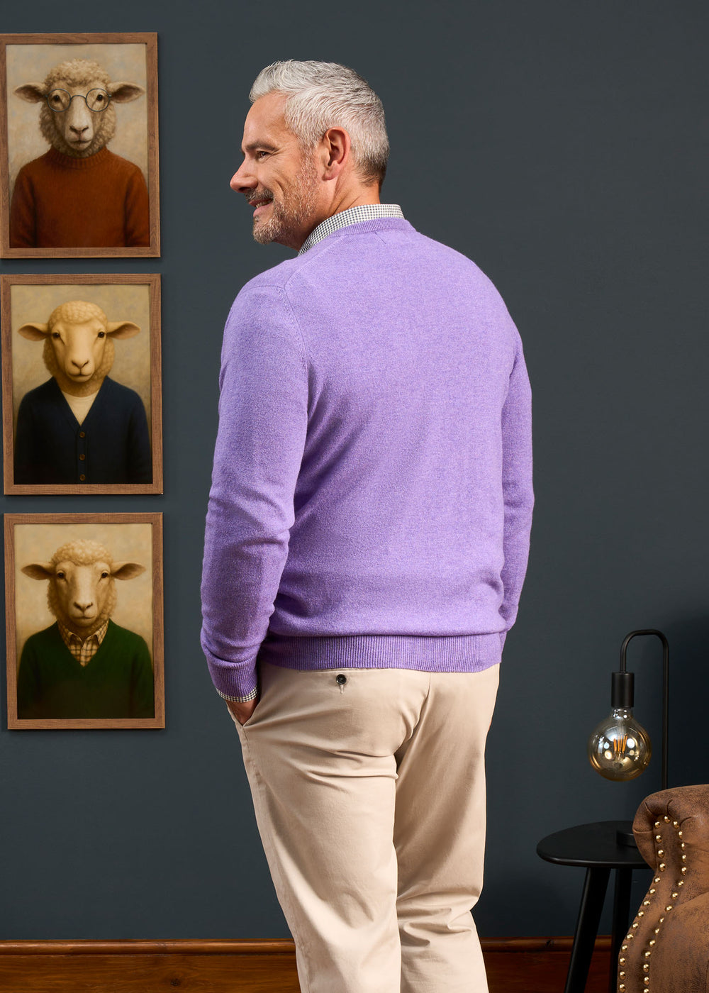 Man with gray hair in Haddington Cashmere Vee Neck Jumper, heliotrope, faces quirky sheep portraits on dark wall.