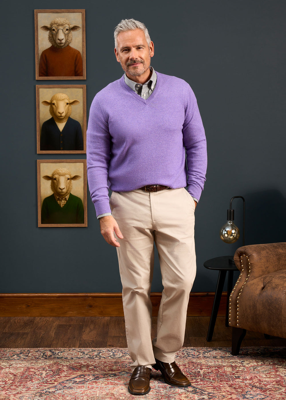 Man wears the Haddington Pure Cashmere Vee Neck Jumper in heliotrope and khakis, with sheep portraits behind him.