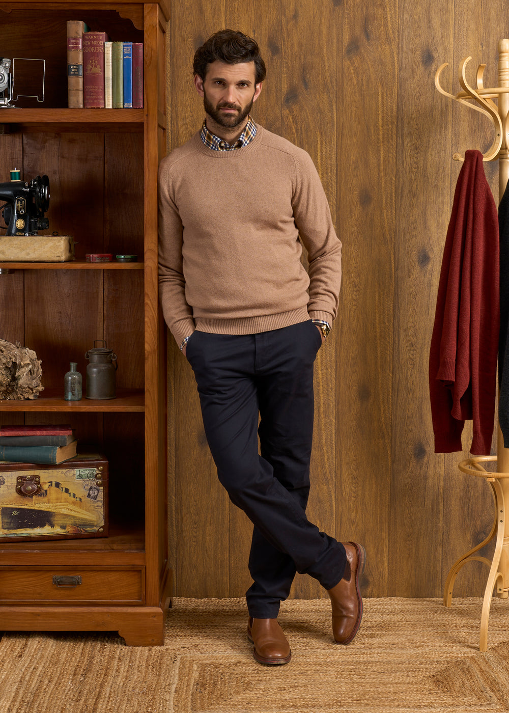 Man in Lenzie Camel Jumper stands by a bookcase in a cozy, wood-paneled room.