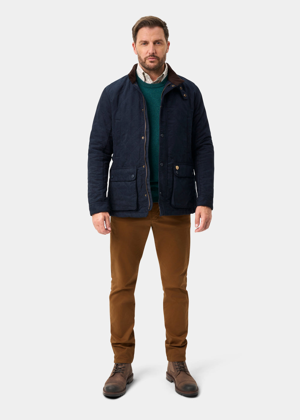 Man in Felwell Men’s Quilted Jacket in Dark Navy, green sweater, brown pants & boots on a plain white background.