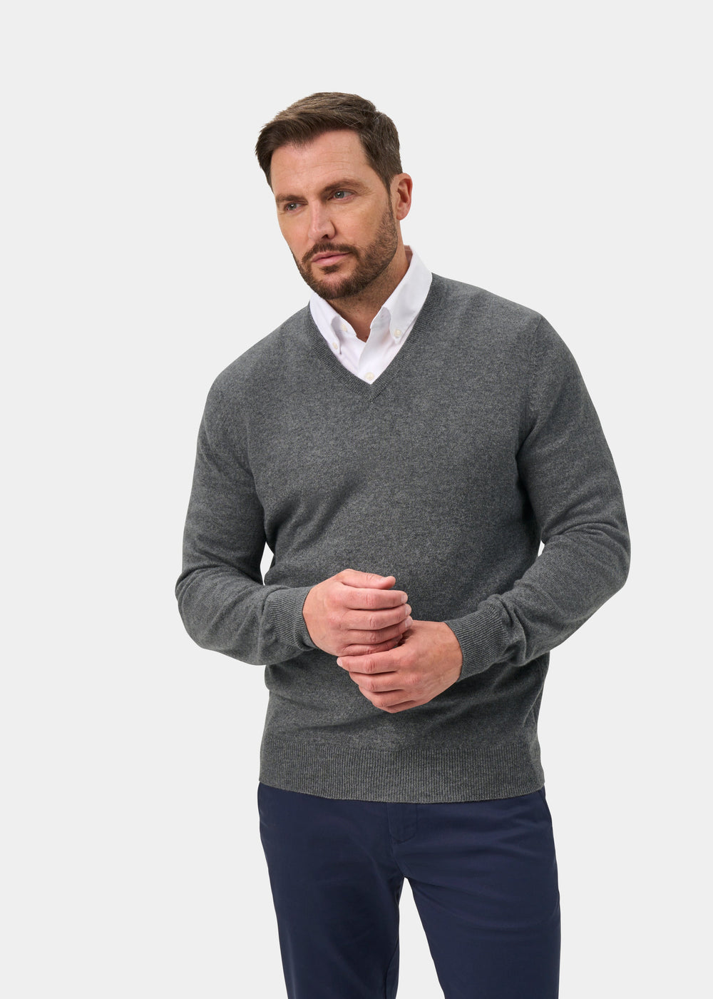 Man wears Haddington Pure Cashmere V-Neck Jumper in Derby over a white shirt and navy pants, against plain background.