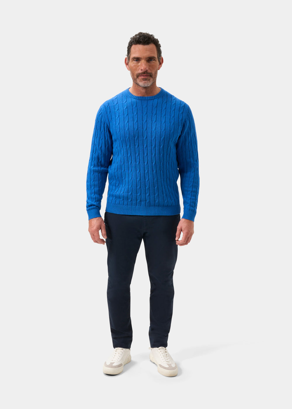 Man wears Honley Regatta cable knit crew neck jumper, dark pants, white sneakers; stands by plain light background.