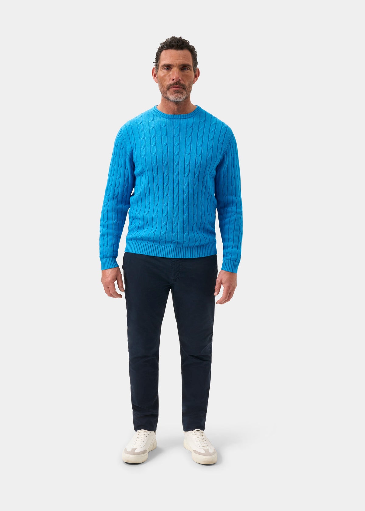 Man in a Honley Blue Lagoon cable knit crew neck jumper, dark pants, and white sneakers on a white background.