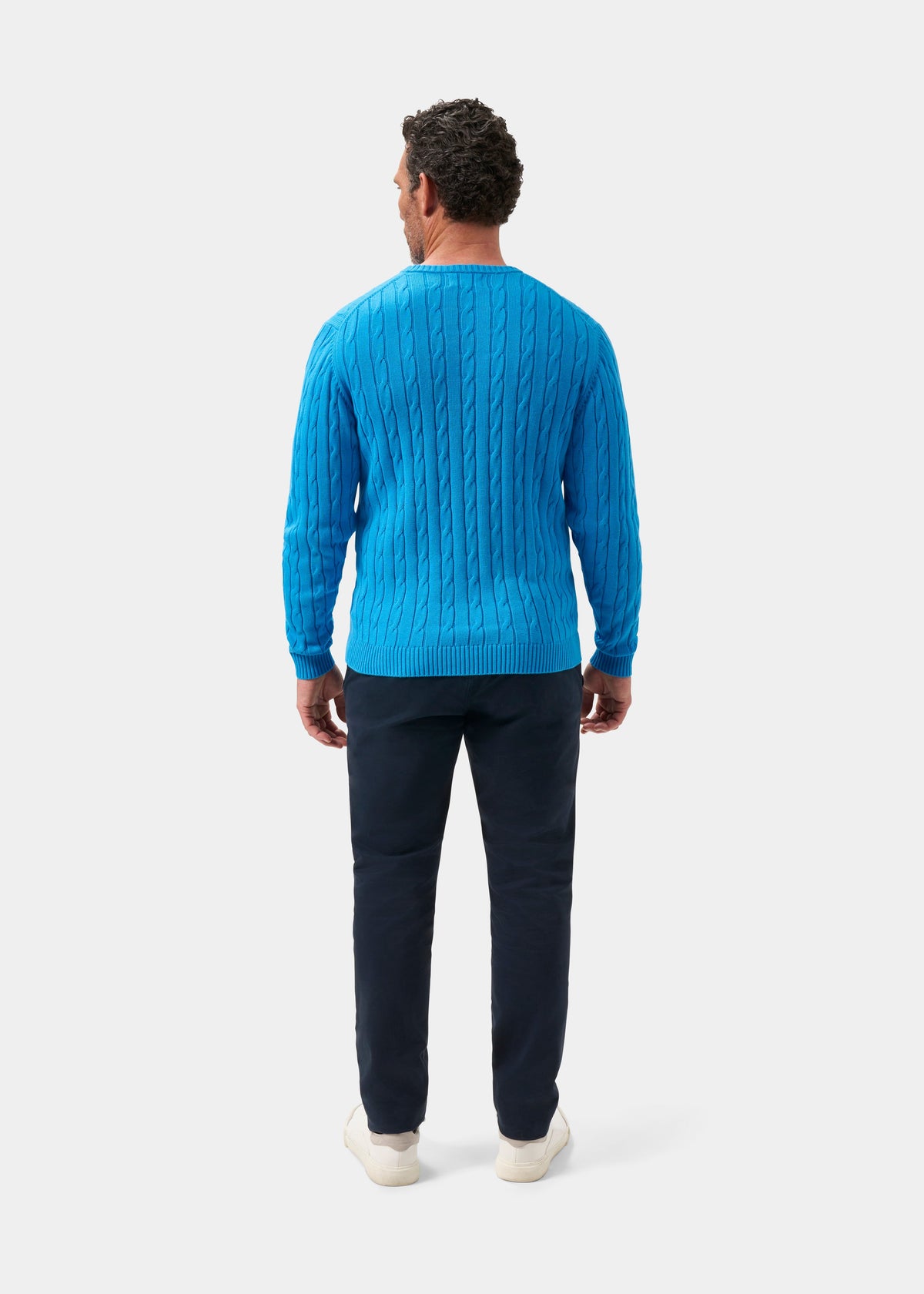 Man facing away in Honley Cotton Cashmere Cable Knit Crew Neck Jumper Blue Lagoon, dark pants, white shoes, plain backdrop.