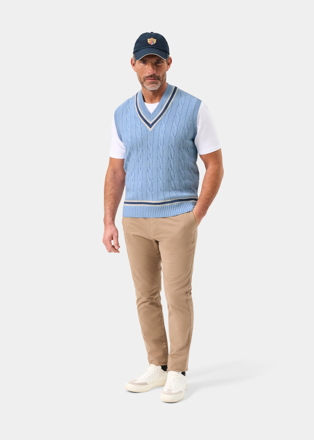 Man wears Chadbury Steel Blue V-Neck Cricket Slipover, white shirt, tan pants, blue cap, and white sneakers.