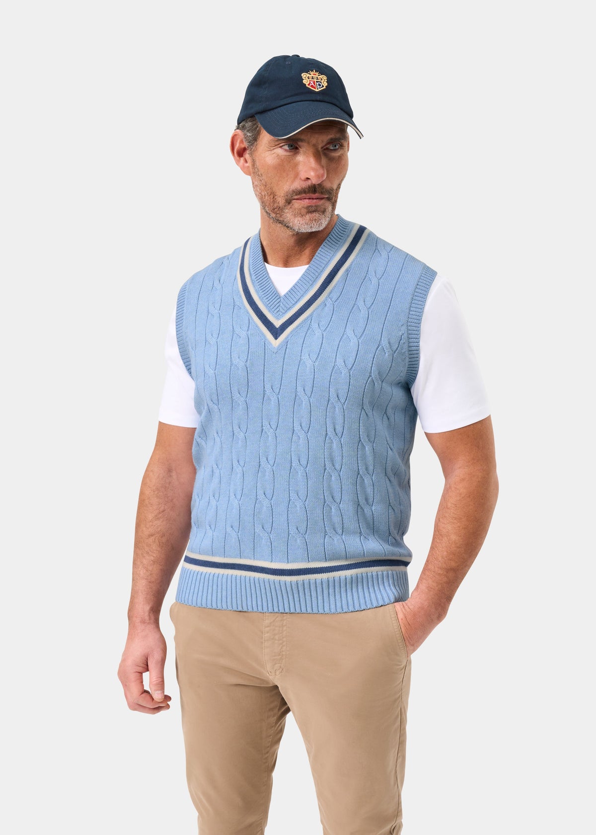 A man in a steel blue Chadbury cotton cable knit slipover, white tee, tan pants, and navy cap stands by a plain backdrop.