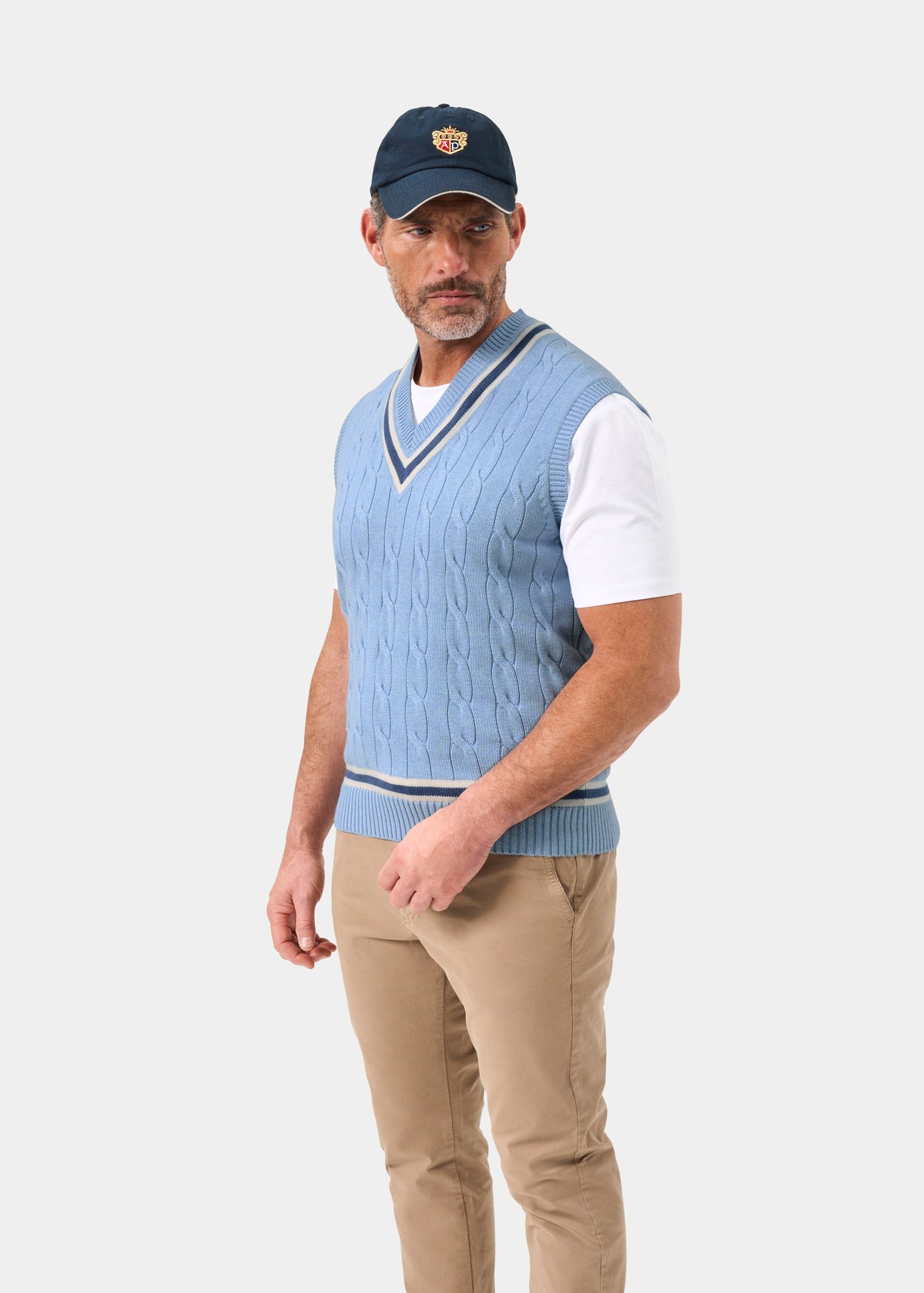 Man in Chadbury cotton cable knit V-neck cricket slipover in steel blue, white tee, beige pants & navy cap stands.
