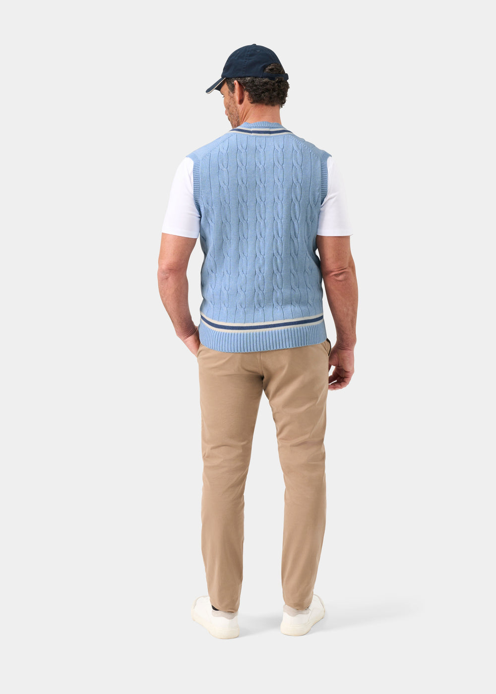 Man in Chadbury Steel Blue V-Neck Cricket Slipover, white shirt, khakis, white shoes, navy cap—back view.