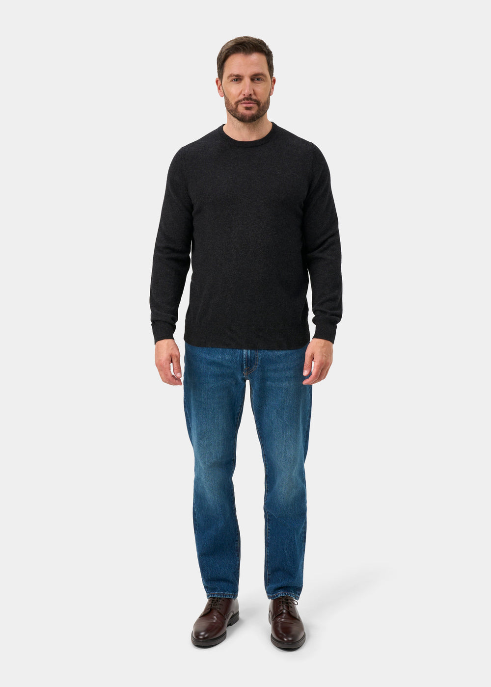 Man wearing a charcoal Brisbane Geelong Wool Crew Neck Jumper, blue jeans, and brown shoes against a white background.