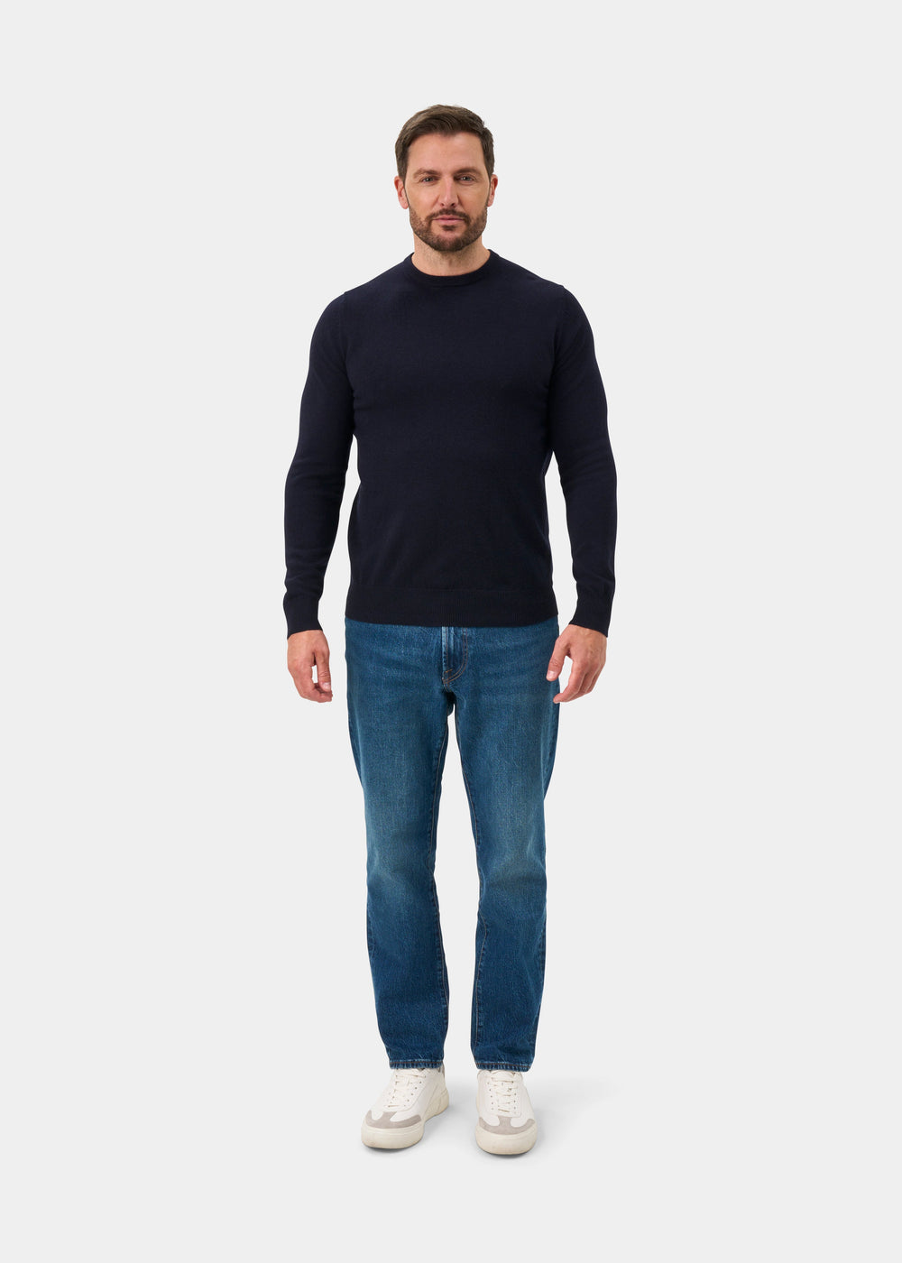 Man stands against a white background in a dark navy Brisbane Geelong Wool Crew Neck Jumper, blue jeans, and sneakers.