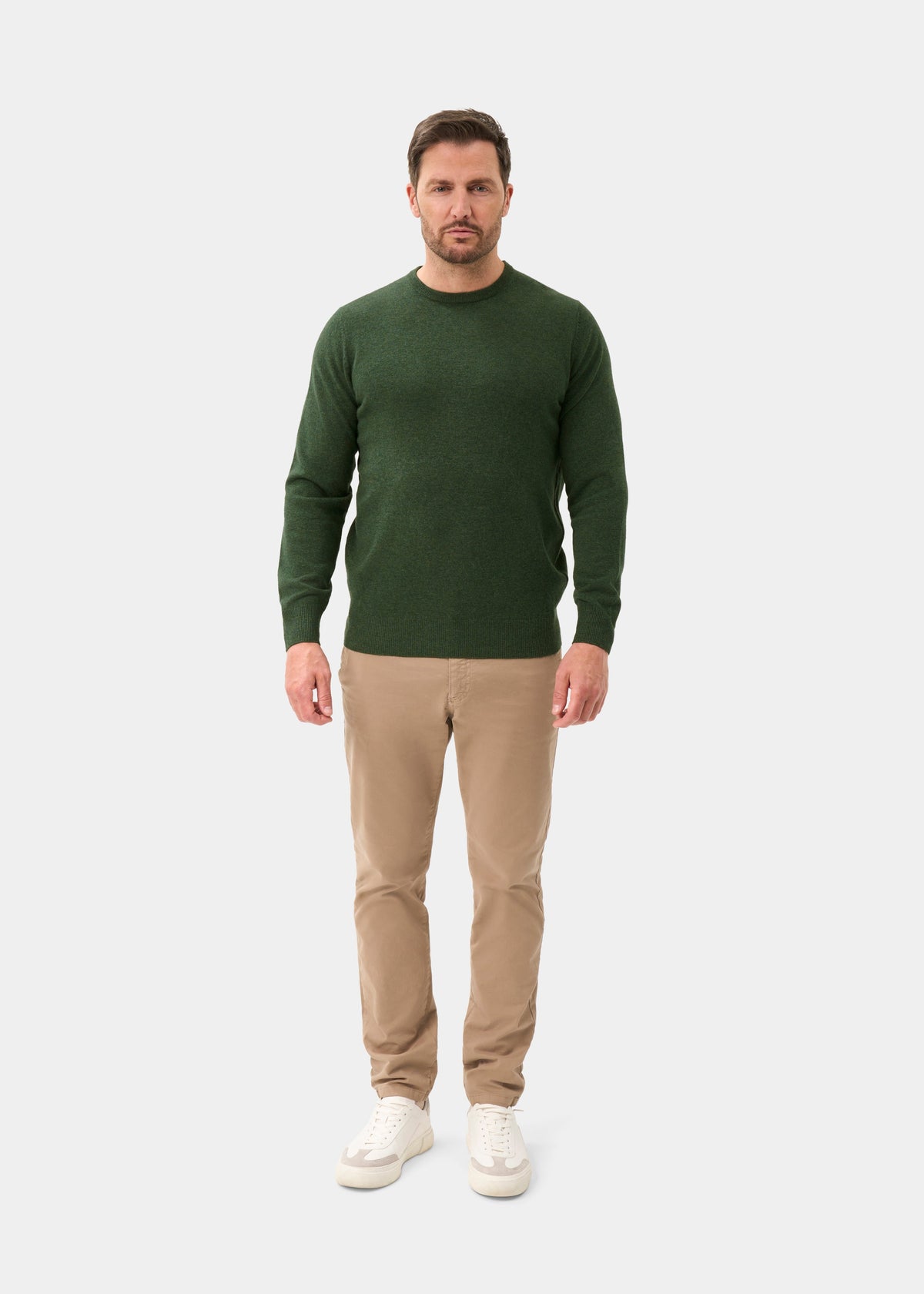 Man in a green Brisbane Geelong Wool Crew Neck Jumper in Rosemary, beige pants, white sneakers, on white background.