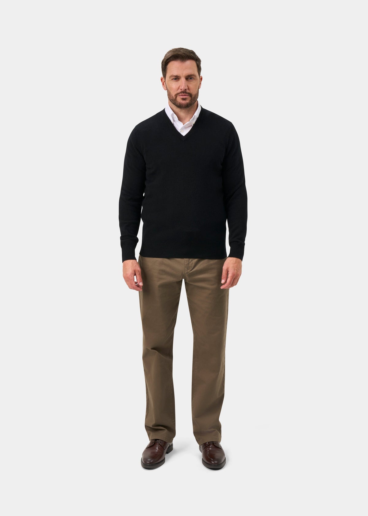 Man in Albury Geelong Wool V-Neck Jumper in Black, white shirt, khaki pants, brown shoes, on plain background.