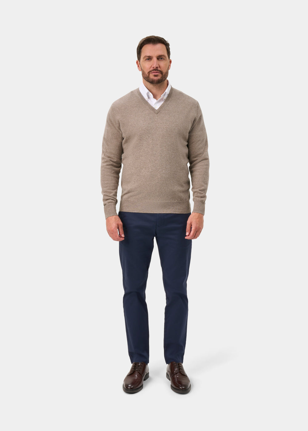 Man wearing Albury Geelong Wool V-Neck Jumper in Mushroom, white shirt, navy pants, and brown shoes on white background.
