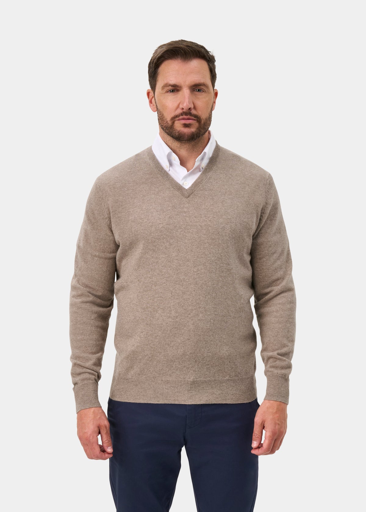 Man in Albury Geelong Wool V-Neck Jumper in Mushroom over a white shirt and navy pants, on a plain white background.