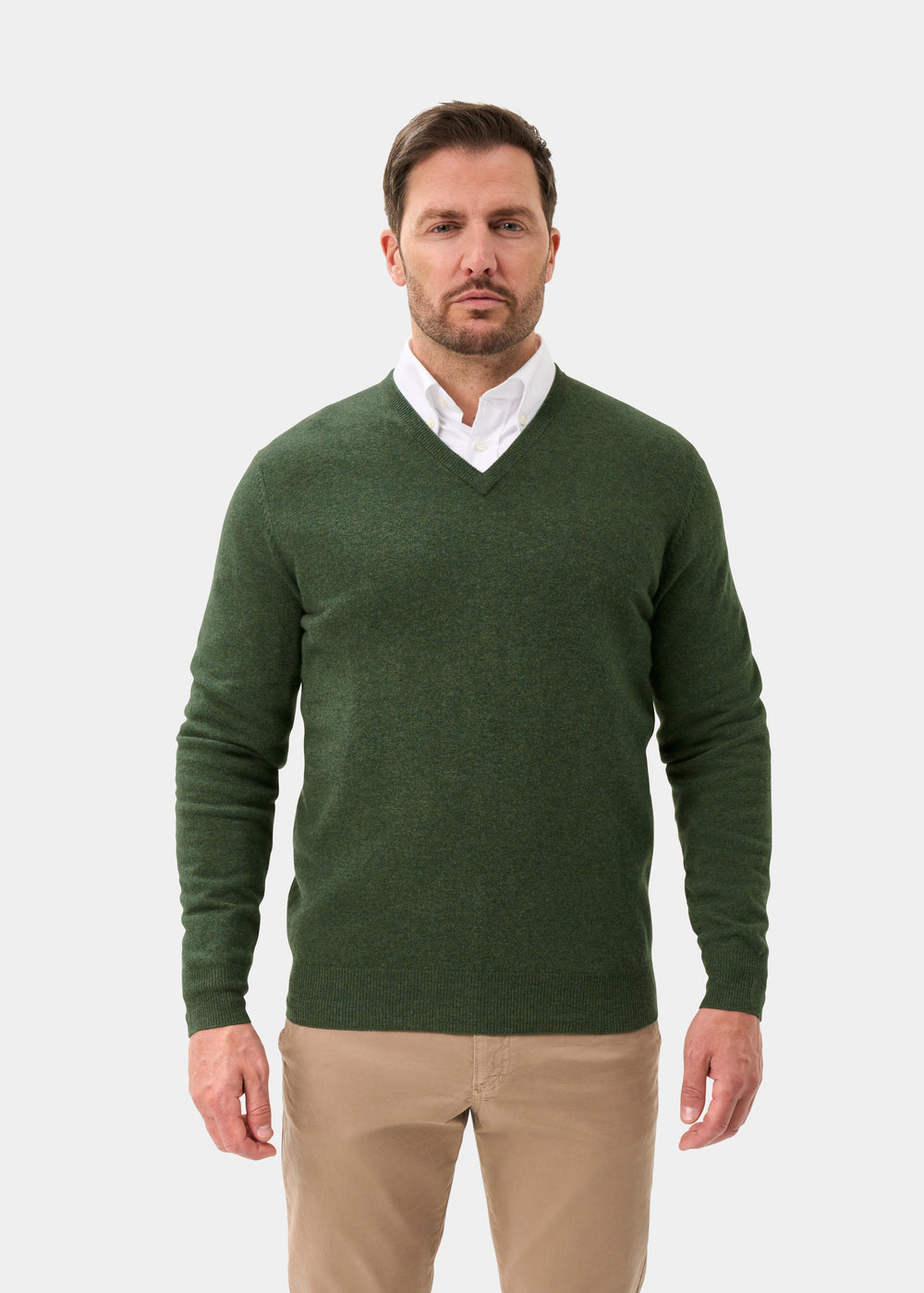 Man in Albury Geelong Wool V-Neck Jumper in Rosemary, over a white shirt and beige pants, against a plain light background.