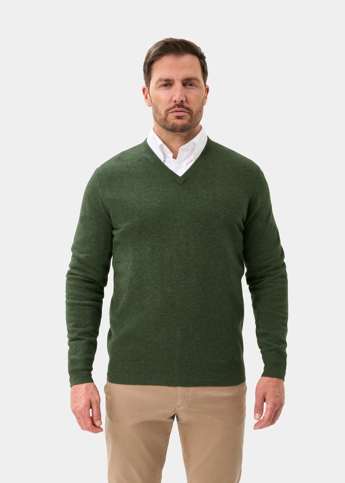 Man in Albury Geelong Wool V-Neck Jumper in Rosemary, over a white shirt and beige pants, against a plain light background.