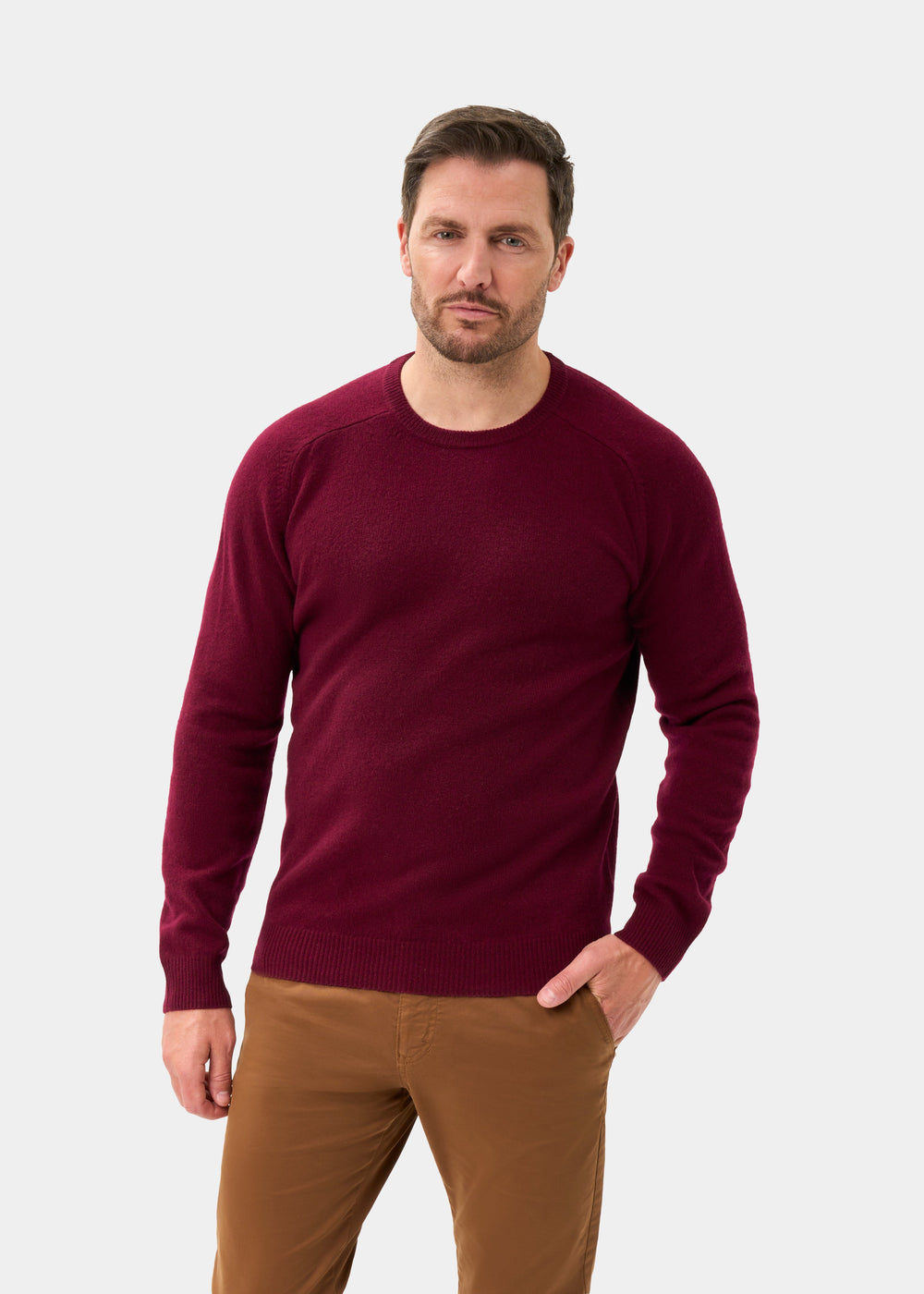 Man wears Lenzie Lambswool Crew Neck Jumper in Bordeaux and brown pants, standing against a plain white background.