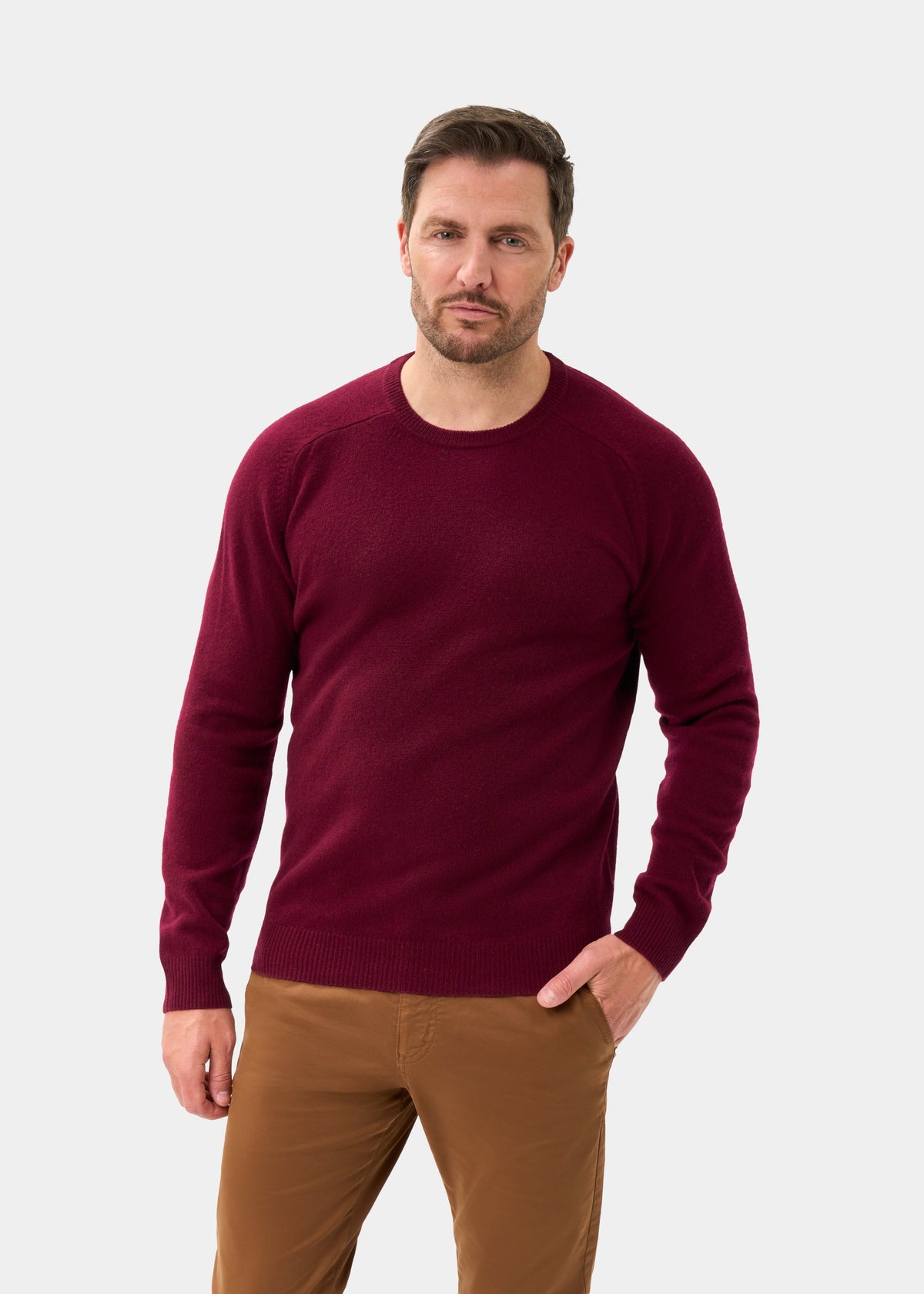 Man wears Lenzie Lambswool Crew Neck Jumper in Bordeaux and brown pants, standing against a plain white background.