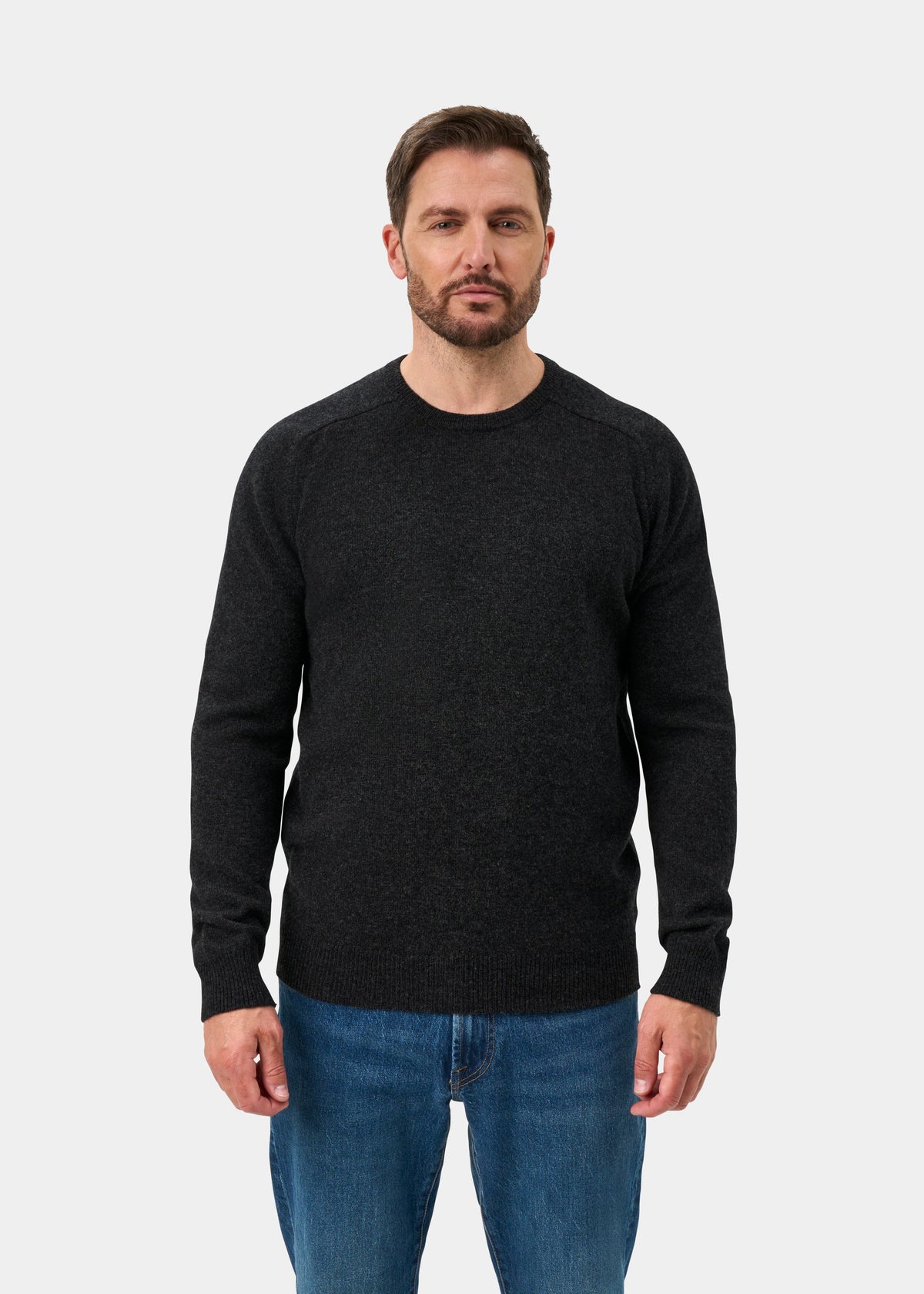 Man wears Lenzie Lambswool Crew Neck Jumper in Charcoal, regular fit, with blue jeans against a white background.