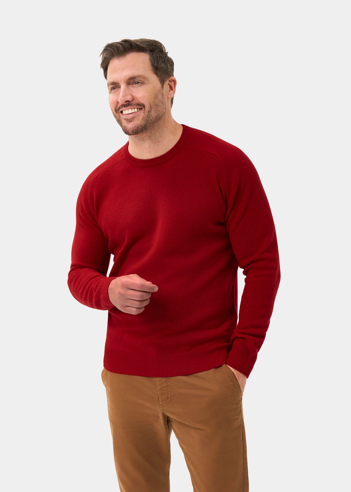 Man wears Lenzie Lambswool Crew Neck Jumper in Dubonnet with tan pants, smiling hand-in-pocket against light backdrop.