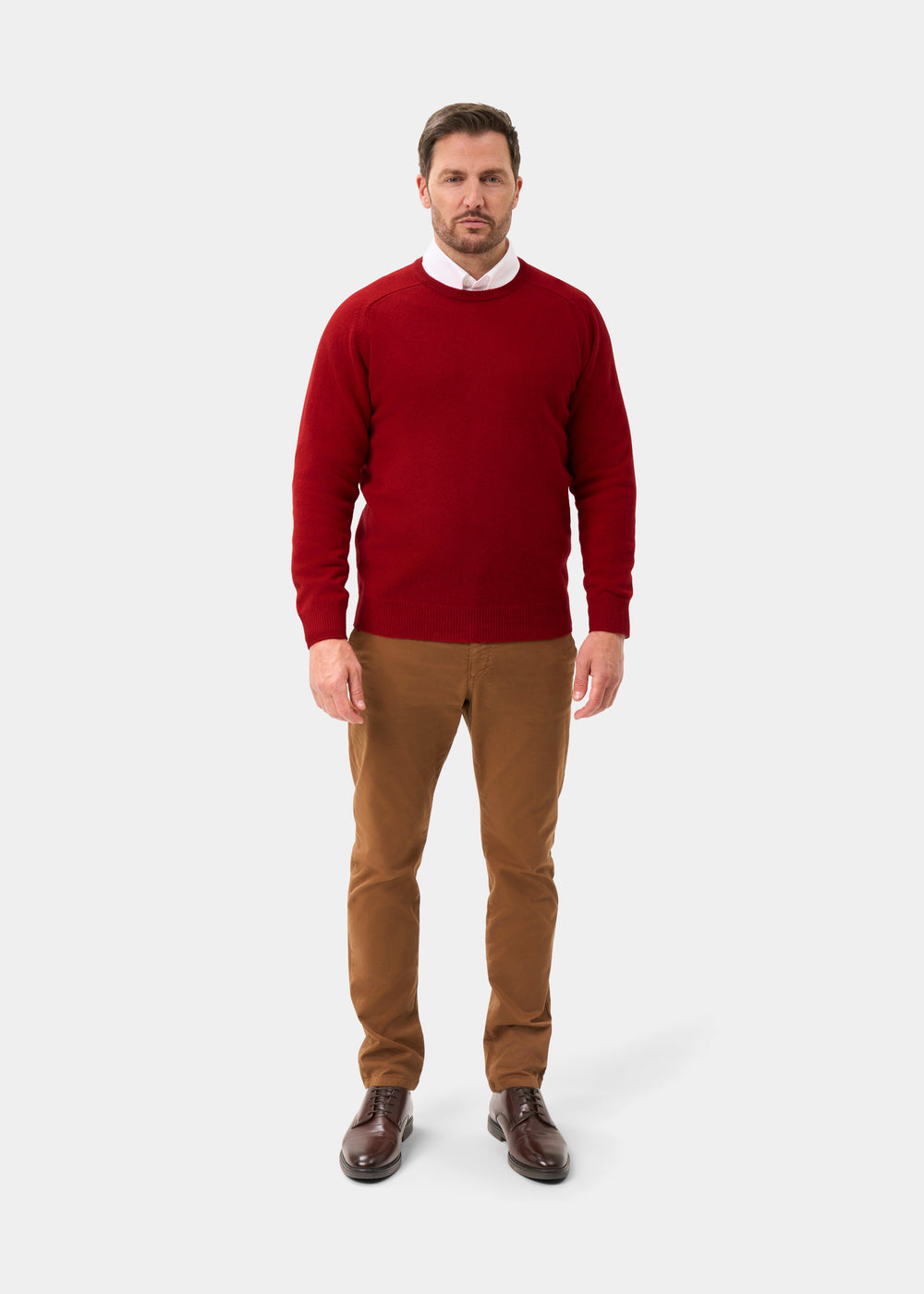Man in Dorset Lambswool Crew Neck Jumper in Dubonnet, white shirt, brown pants and shoes on a plain light background.