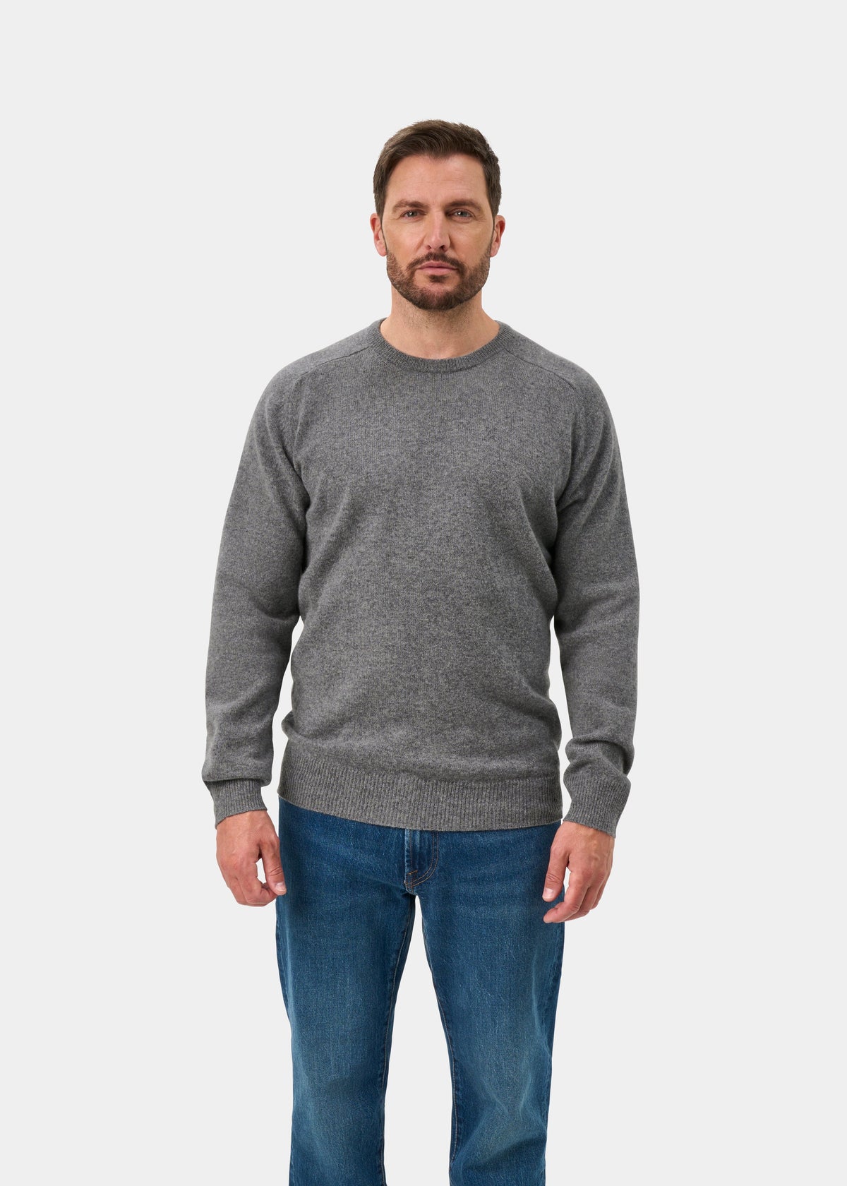 Man wears Dorset Lambswool Crew Neck Jumper in Grey Mix, classic fit, with blue jeans, facing forward on white background.