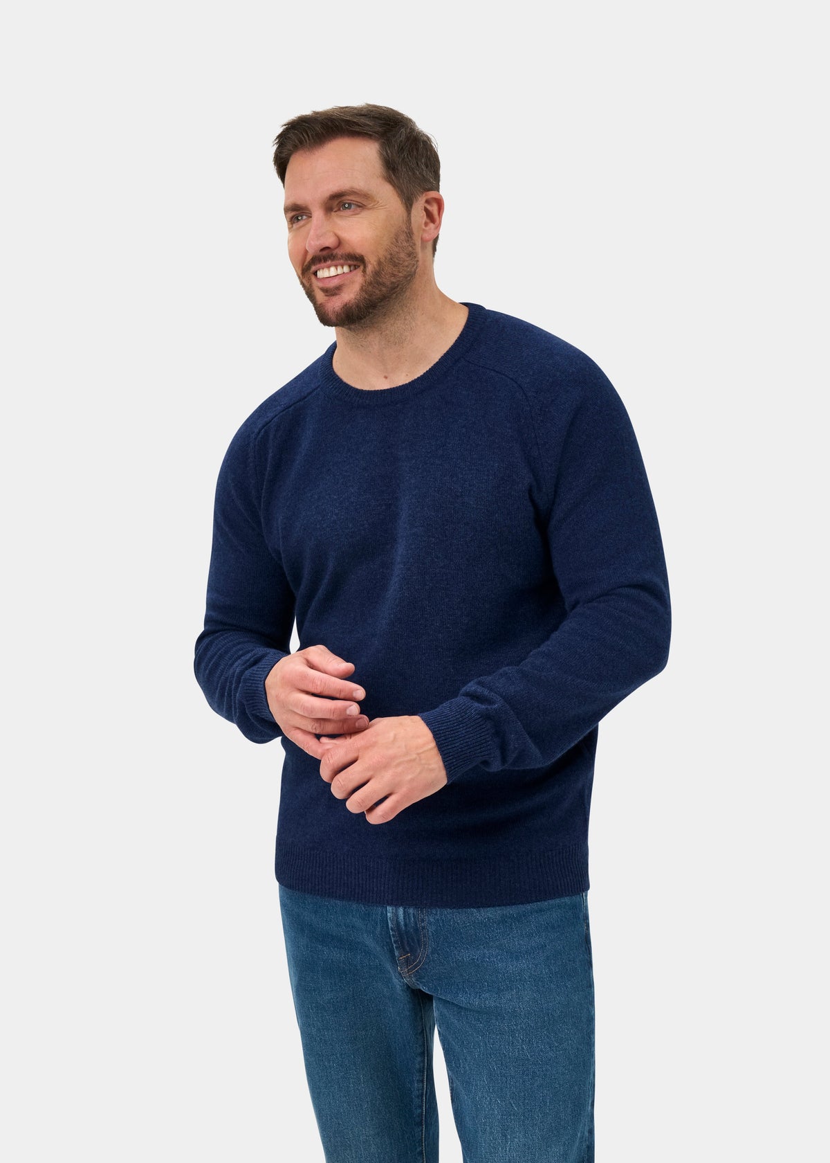 Smiling man wears the Lenzie Lambswool Crew Neck Jumper in Rhapsody with blue jeans, standing by a plain gray backdrop.