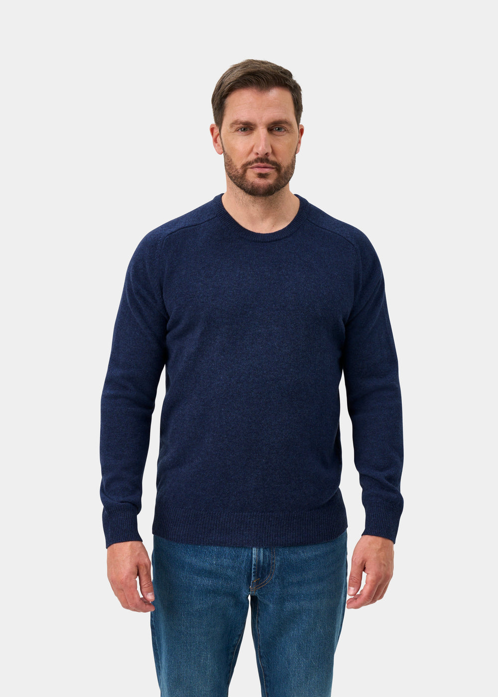 A man wears the Dorset Lambswool Crew Neck Jumper in Rhapsody, classic fit, with blue jeans against a light gray backdrop.
