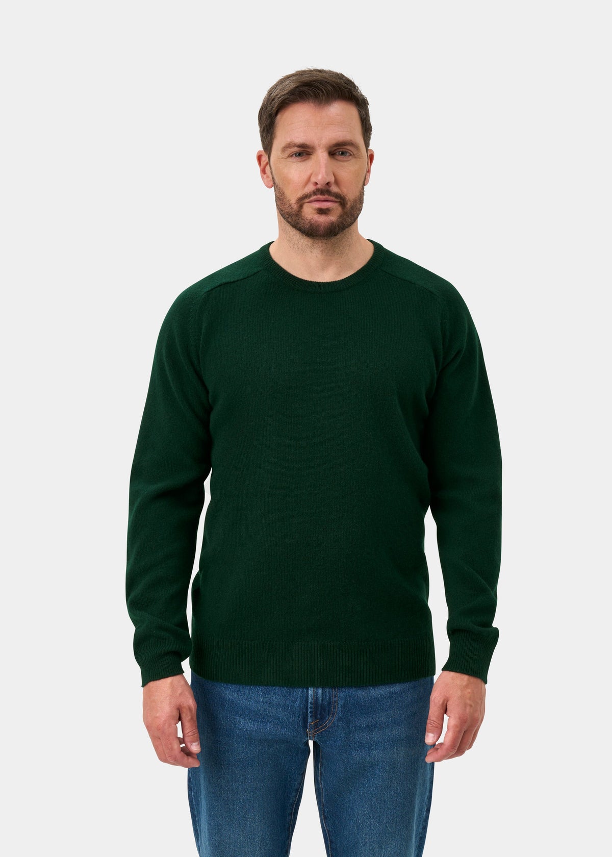 Man wears Dorset Lambswool Crew Neck Jumper in Tartan Green, classic fit, with blue jeans against a plain background.