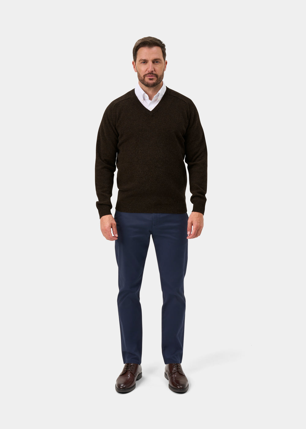 Man in Kilsyth Lambswool Vee Neck Jumper in Cocoa, white shirt, blue pants, brown shoes stands against a white background.