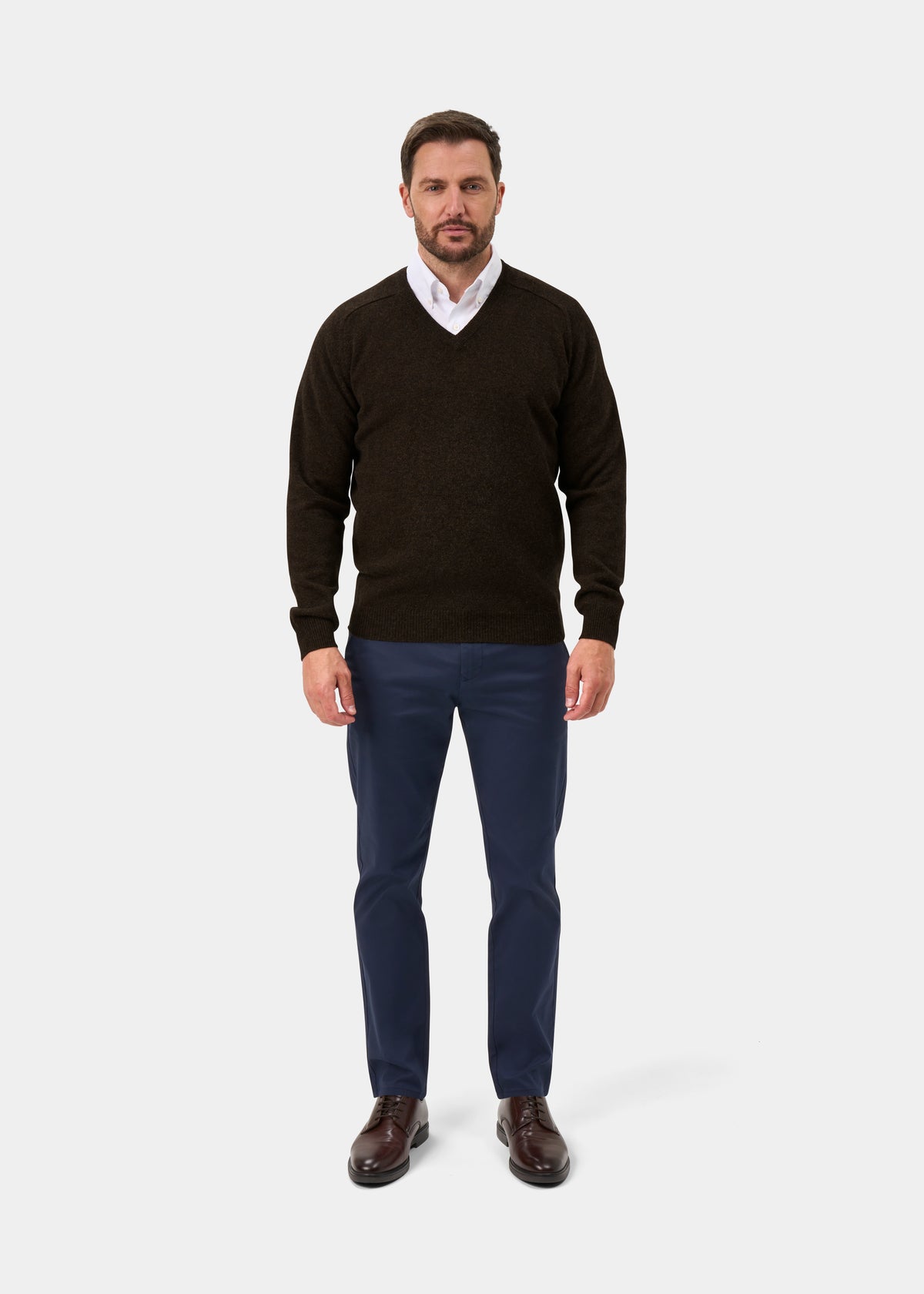 Man in Kilsyth Lambswool Vee Neck Jumper in Cocoa, white shirt, blue pants, brown shoes stands against a white background.