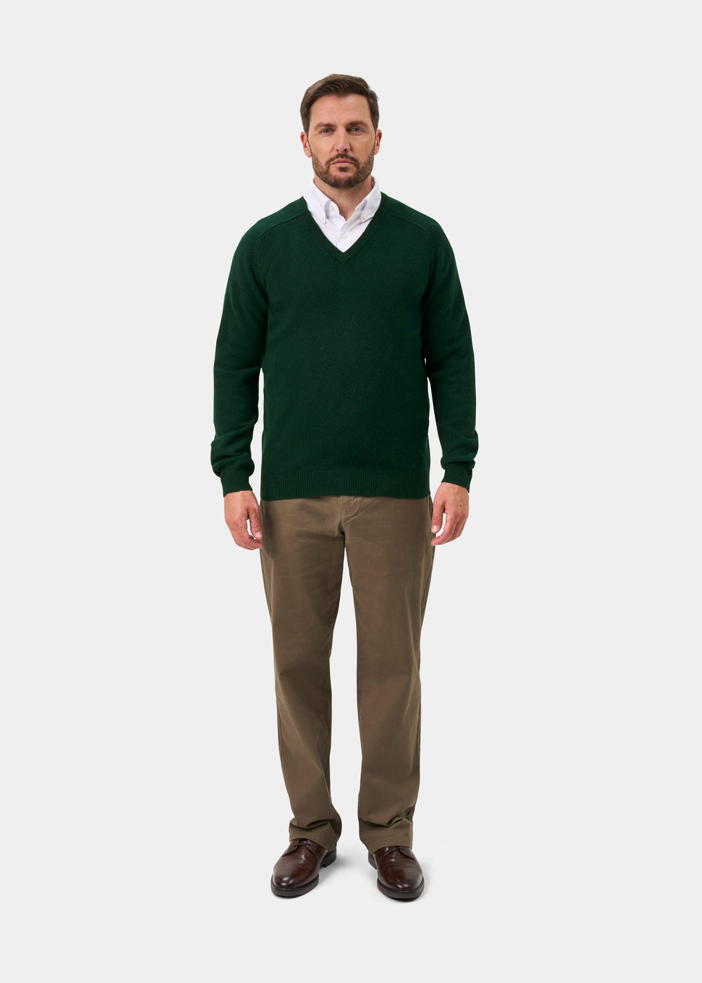 Man in Hampshire Lambswool V-Neck Jumper in Tartan Green, white shirt, khakis, and brown shoes stands against backdrop.