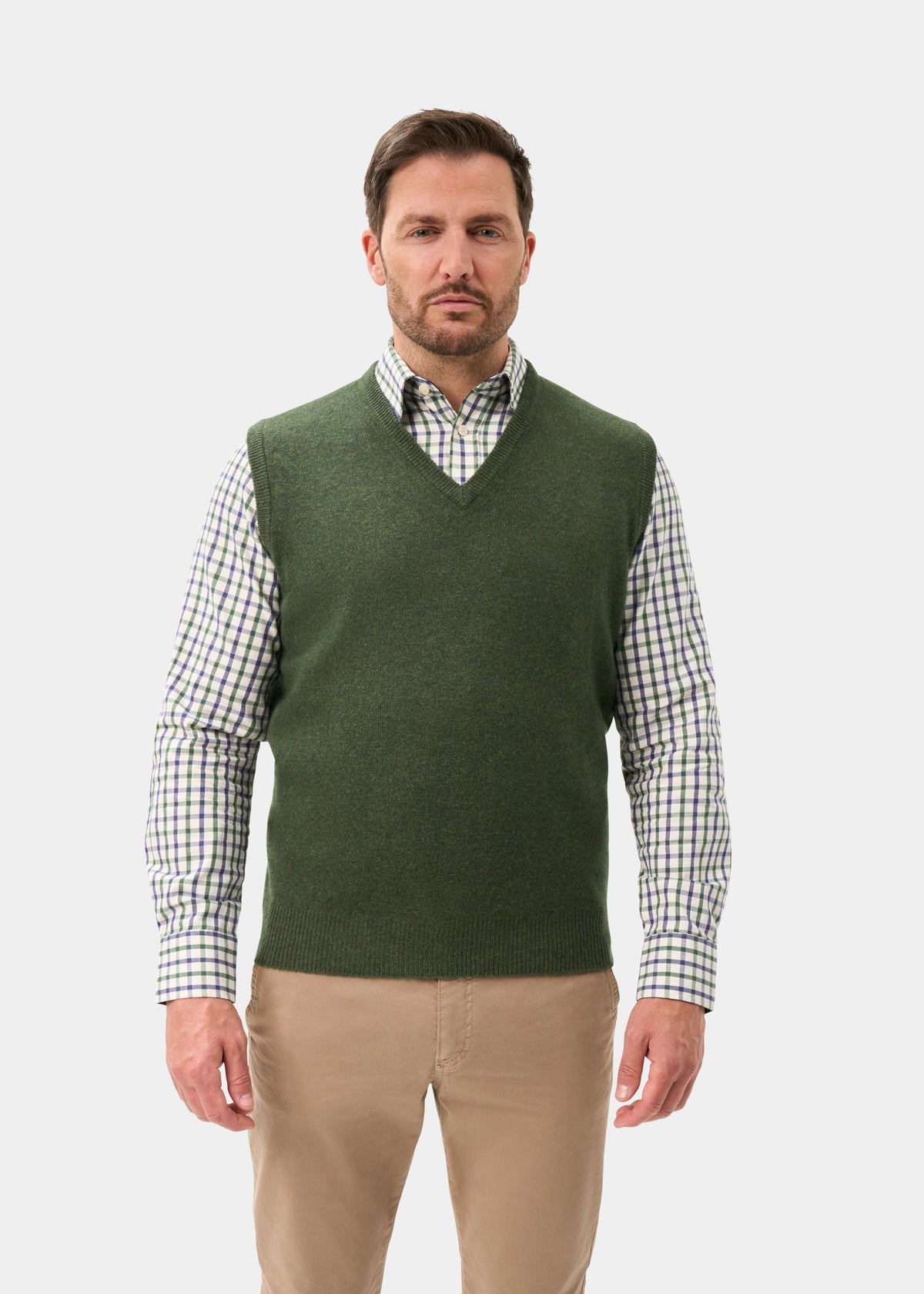 A man wearing a Norfolk Lambswool V-Neck Slipover In Rosemary over a plaid button-down shirt and tan trousers.