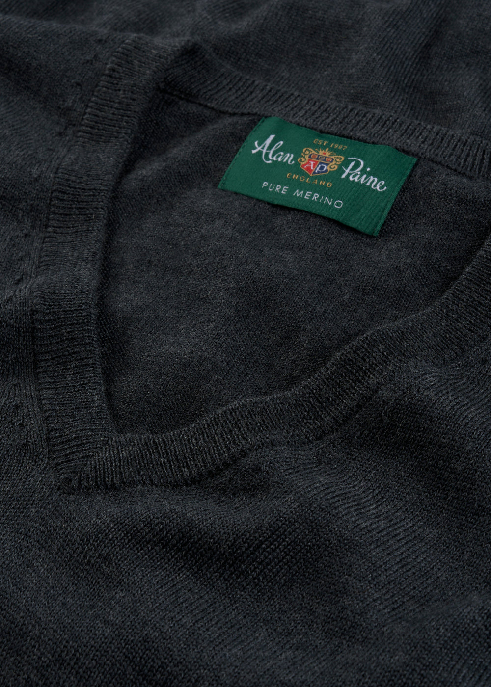 Close-up of a dark charcoal gray knit sweater showing the ribbed v-neck collar and a green woven brand label that reads 'Alain Paine Pure Merino'.