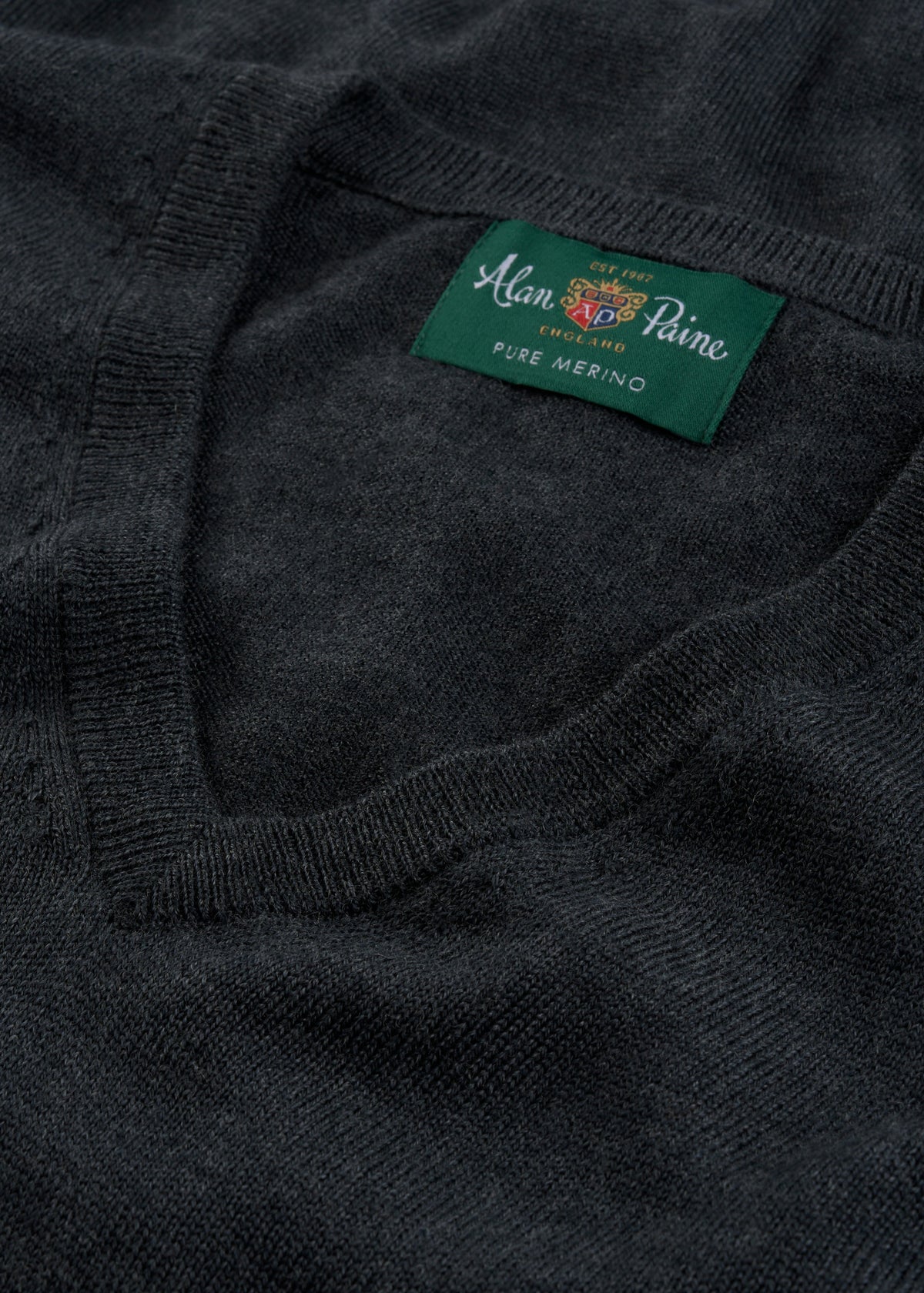 Close-up of a dark charcoal gray knit sweater showing the ribbed v-neck collar and a green woven brand label that reads 'Alain Paine Pure Merino'.