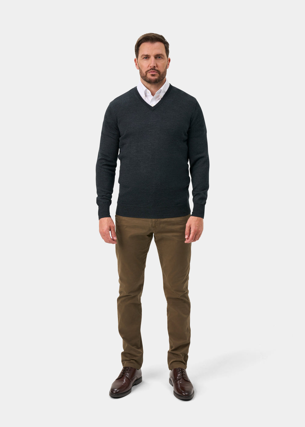 A man wearing a dark charcoal gray v-neck sweater over a white collared shirt, paired with brown trousers and dark brown leather dress shoes, standing against a light gray studio background.