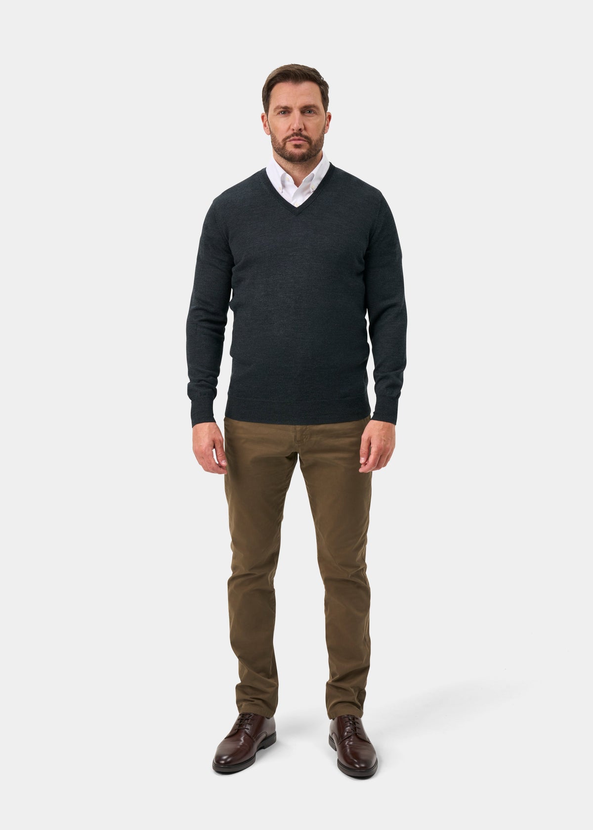 A man wearing a dark charcoal gray v-neck sweater over a white collared shirt, paired with brown trousers and dark brown leather dress shoes, standing against a light gray studio background.