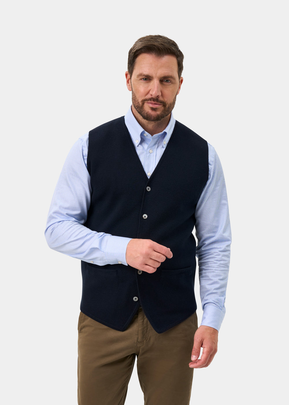 A man wears a Thornley Merino Wool Tailored Dark Navy Waistcoat over a light blue striped dress shirt and brown trousers.