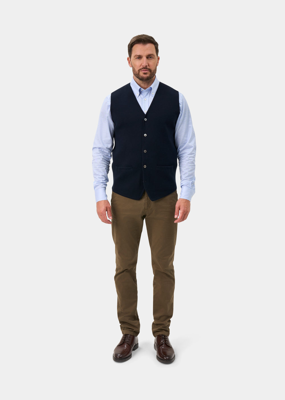 A man wears a Thornley Merino Wool Tailored Dark Navy Waistcoat over a light blue shirt, brown trousers, and dark shoes.
