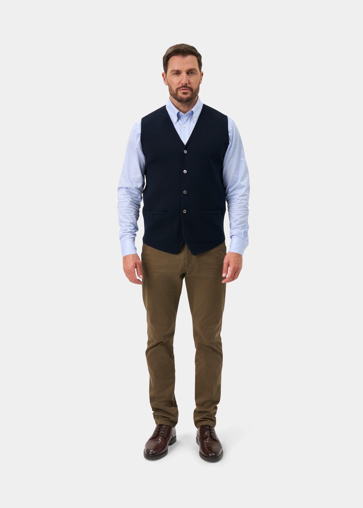 A man wears a Thornley Merino Wool Tailored Dark Navy Waistcoat over a light blue shirt, brown trousers, and dark shoes.