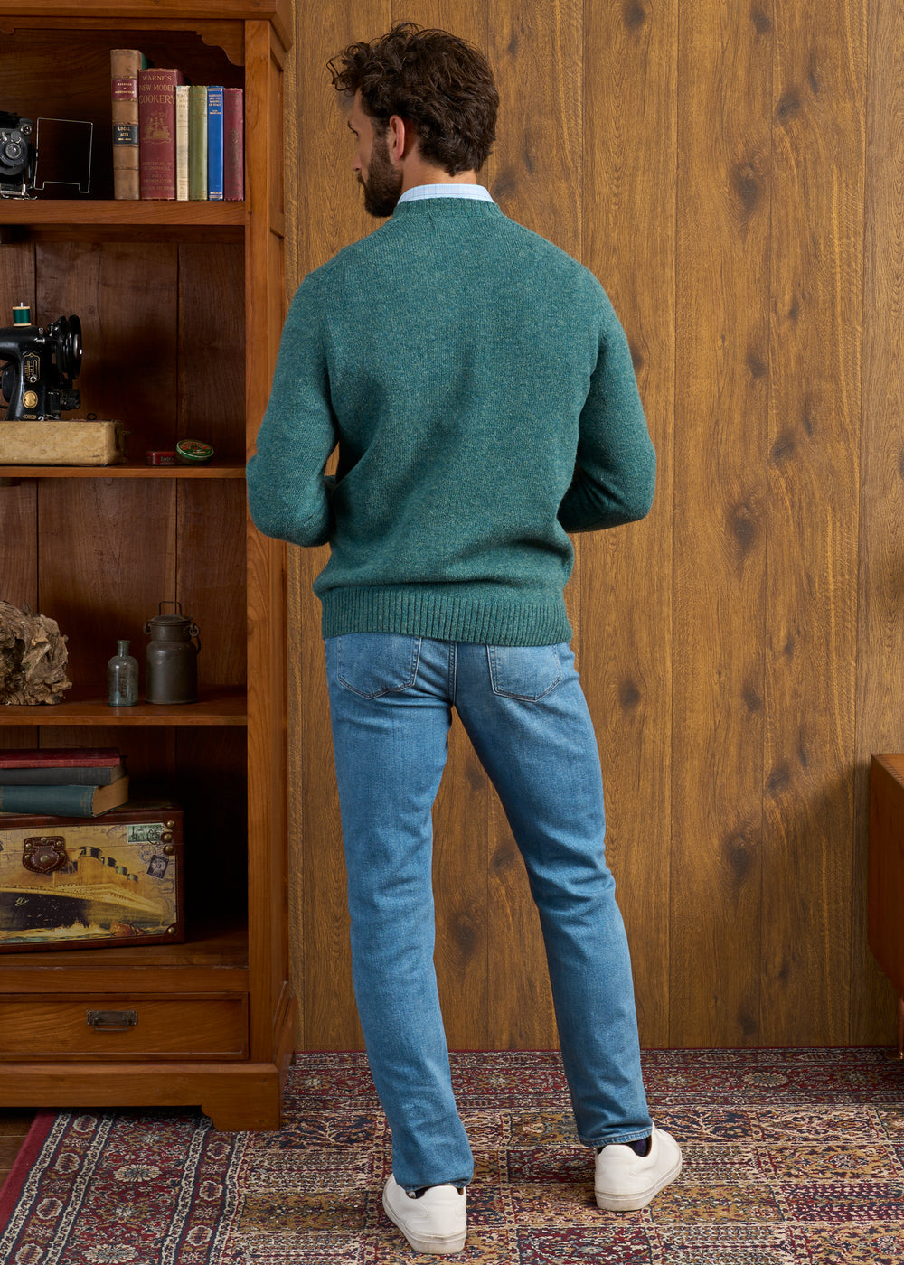 Man in a Kingfisher Kinnadie Supersoft Shetland Wool Crew Neck Jumper and jeans stands in a cozy, wood-paneled room.