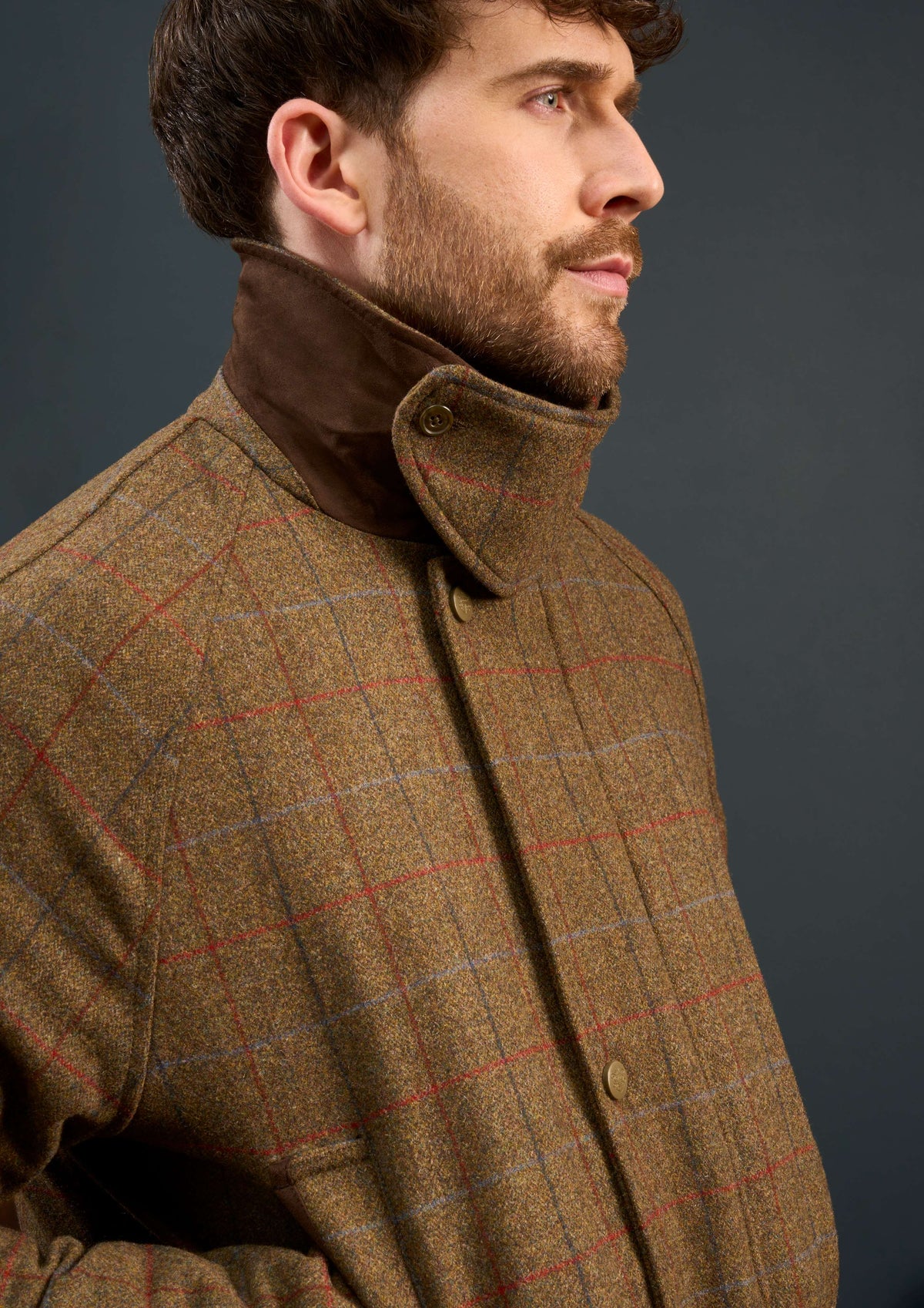Man wearing the Surrey Men’s Tweed Waterproof Coat in Hawkswood, high collar up, gazes sideways against a dark backdrop.