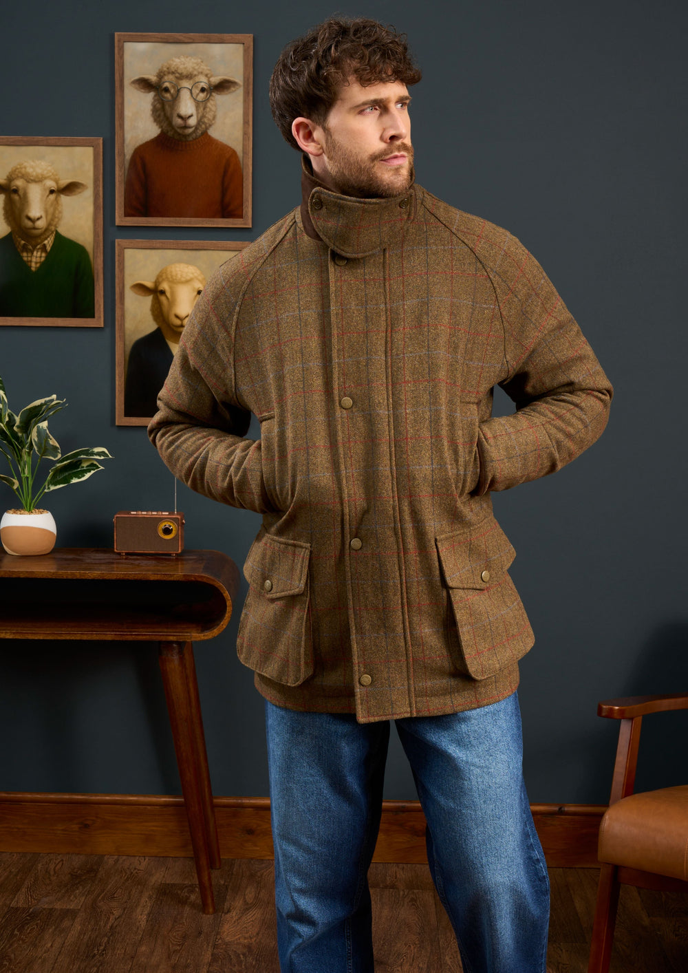 A man in a Surrey Men’s Tweed Waterproof Coat (Hawkswood, Shooting Fit) stands before sheep portraits on the wall.