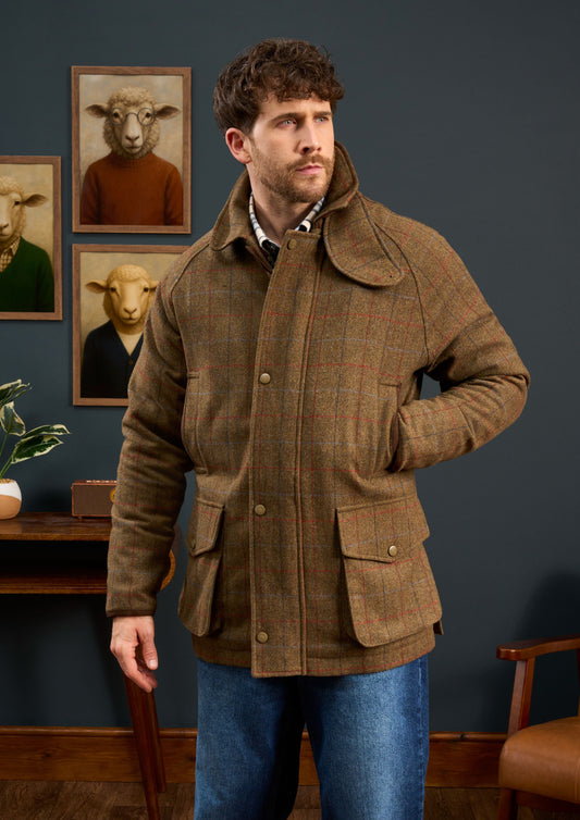 Man in a Surrey Men’s Tweed Waterproof Coat in Hawkswood stands before sheep portraits on a dark wall.