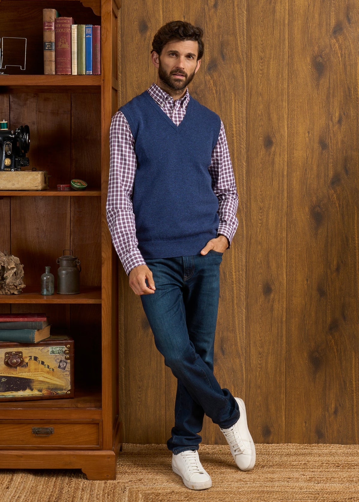 Man in a Norfolk Lambswool V-Neck Slipover In Rhapsody worn over a plaid shirt and jeans, leaning against a wooden bookshelf.