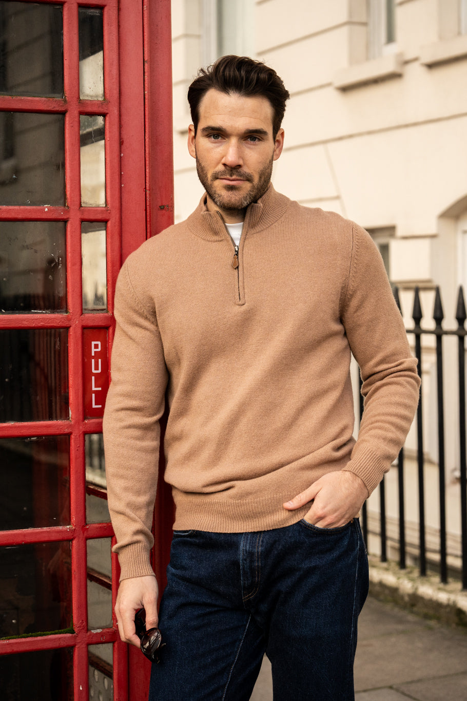 A man in a Sutherland Lambswool Quarter Zip Jumper In Camel stands next to a red telephone box.