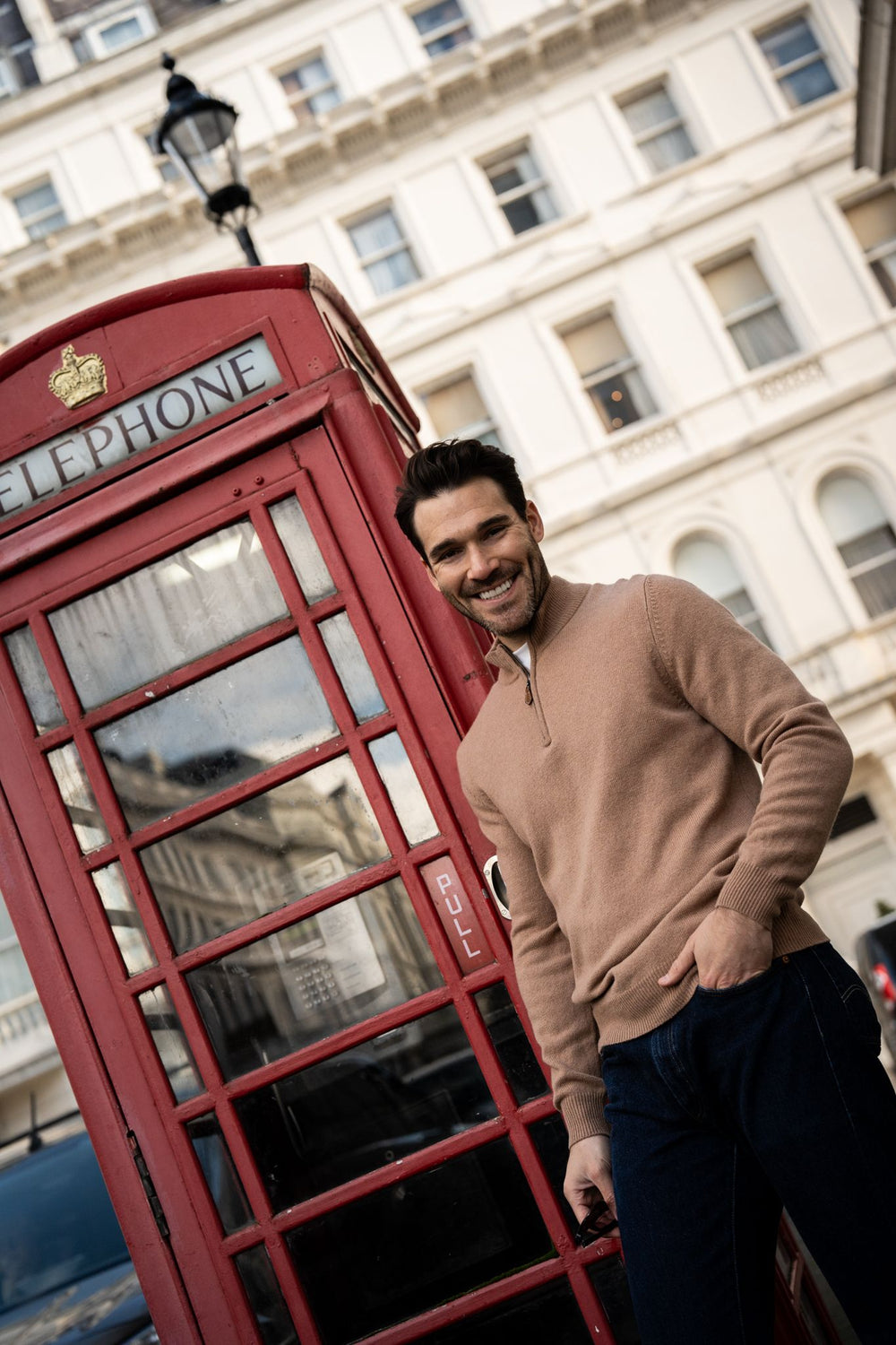 Smiling man in a Sutherland Lambswool Quarter Zip Jumper In Camel standing next to a red British telephone box.