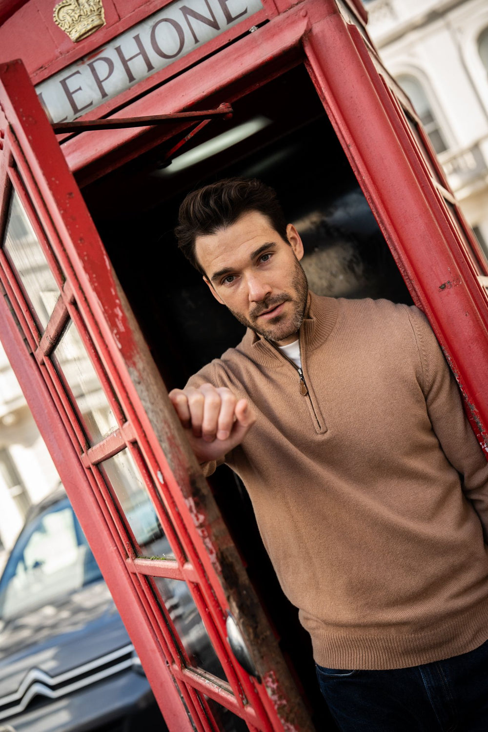 A man in a Sutherland Lambswool Quarter Zip Jumper In Camel stands inside a red telephone booth.